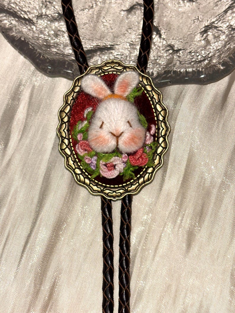May include: A bolo tie featuring an oval antique bronze frame containing a needle-felted white rabbit head surrounded by embroidered pink and green flowers on a deep red background, attached to a braided dark brown leather cord.