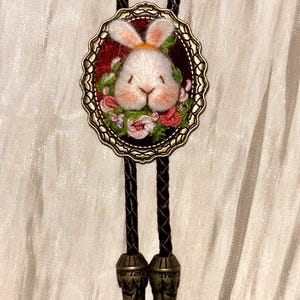 May include: A bolo tie featuring a central oval bronze-toned frame containing an embroidered white rabbit head surrounded by small pink and green flowers on a dark red background. The cord is dark brown braided leather with metal tips.