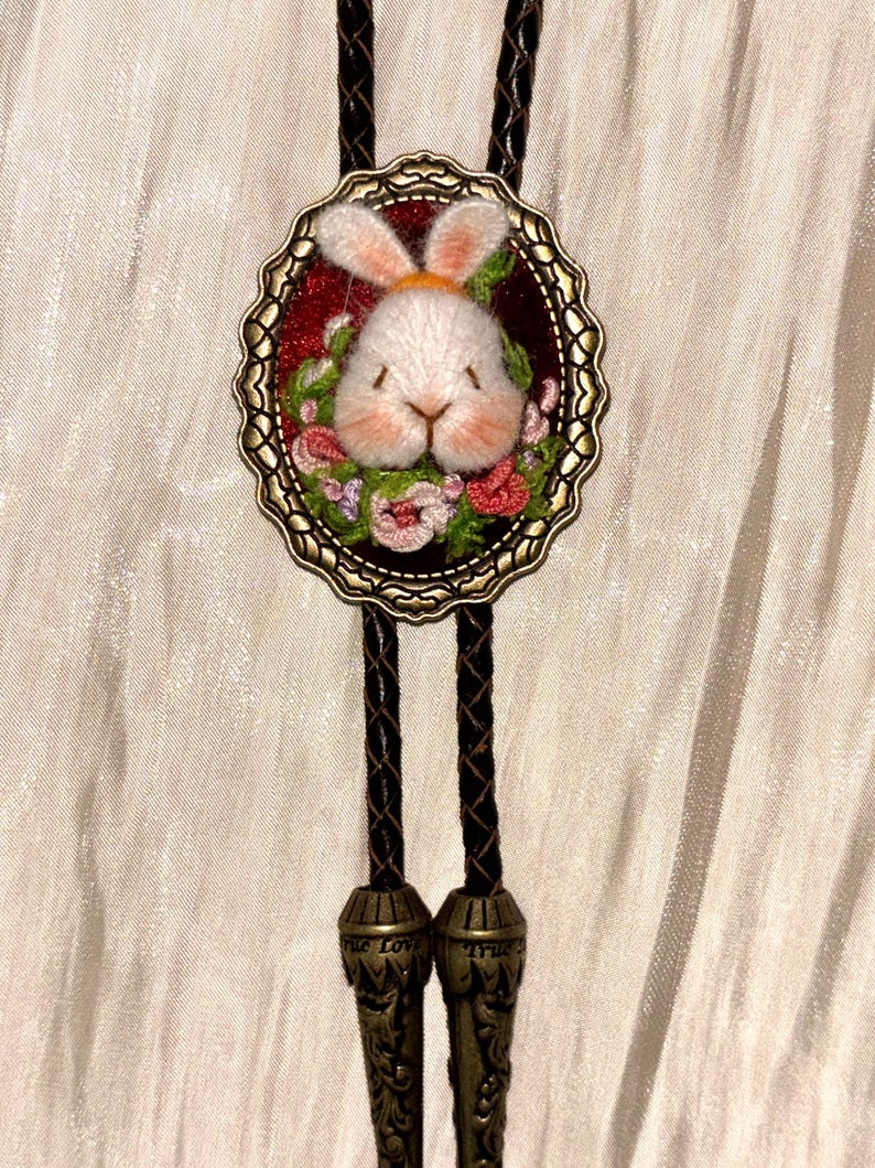 May include: A bolo tie featuring a central oval bronze-toned frame containing an embroidered white rabbit head surrounded by small pink and green flowers on a dark red background. The cord is dark brown braided leather with metal tips.