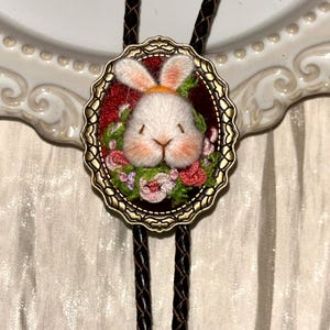 May include: A bolo tie featuring an oval antique brass frame with a scalloped edge. The center displays a white embroidered rabbit head with pink accents, surrounded by small pink and green floral embroidery on a deep red velvet background.