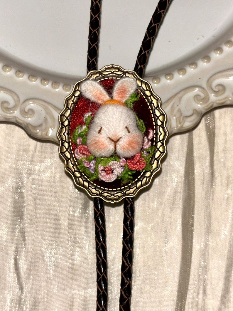 May include: A bolo tie featuring an oval antique brass frame with a scalloped edge. The center displays a white embroidered rabbit head with pink accents, surrounded by small pink and green floral embroidery on a deep red velvet background.