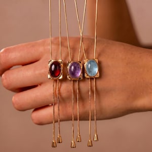 May include: Three gold-toned necklaces featuring thin snake chains and rectangular pendants. Each pendant holds a smooth, oval-shaped cabochon stone in deep red, light purple, and pale blue, with two dangling chain ends finished with small metal caps.