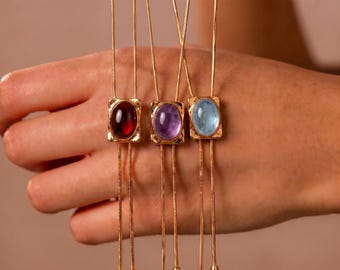 May include: Three gold-toned necklaces featuring thin snake chains and rectangular pendants. Each pendant holds a smooth, oval-shaped cabochon stone in deep red, light purple, and pale blue, with two dangling chain ends finished with small metal caps.