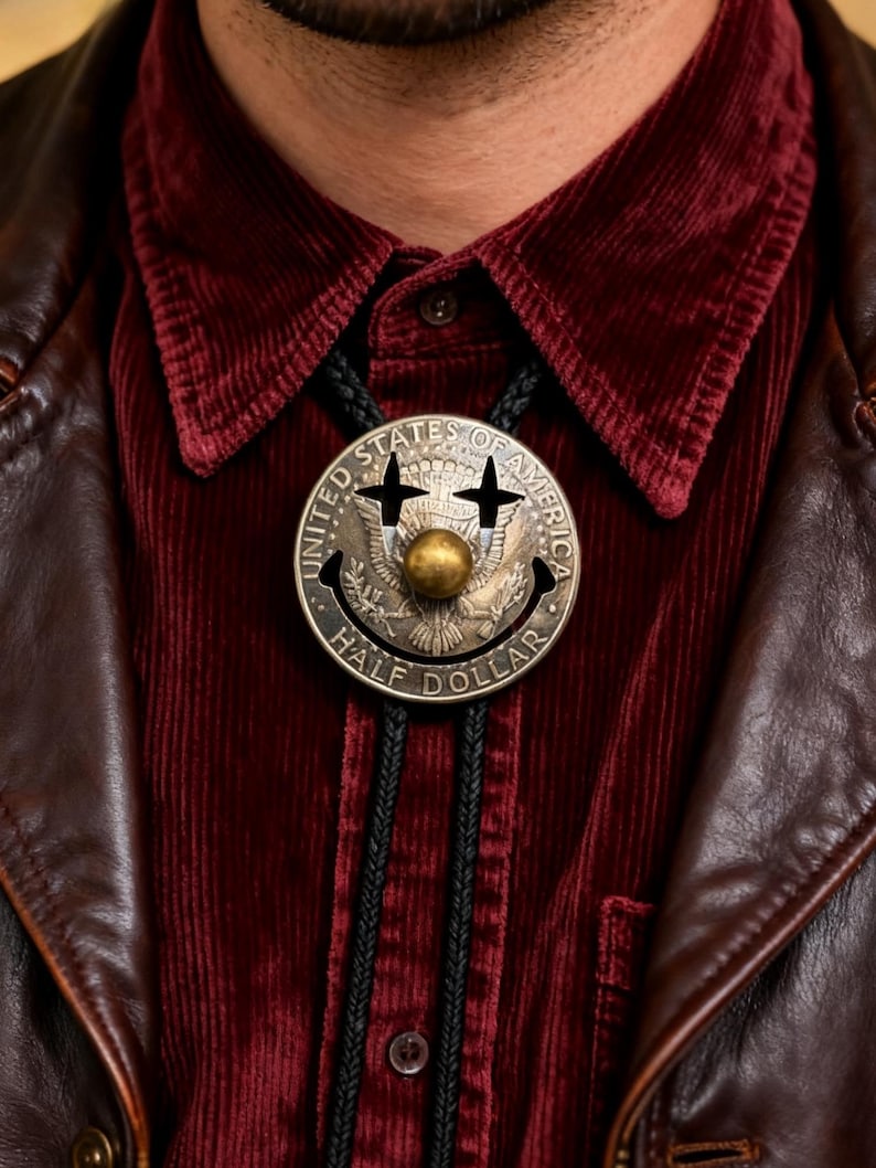 May include: A silver-toned bolo tie featuring a smiling face design with a gold-colored ball in the center. The tie is made from a United States half dollar coin. The bolo tie is worn over a burgundy corduroy shirt and a brown leather jacket.