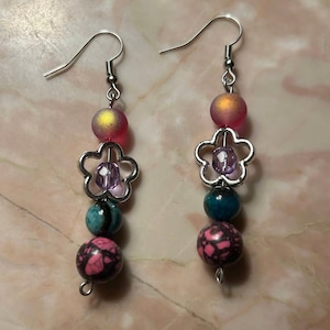 May include: A pair of dangle earrings with a silver flower charm, pink and blue patterned beads, and a purple bead in the center of the flower.