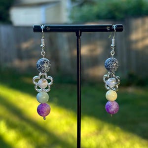 Puede incluir: Pendientes colgantes con diseño floral. Cada pendiente presenta un dije de flor plateada, una cuenta con estampado blanco y negro, una cuenta verde claro, una cuenta amarilla y una cuenta morada. Los pendientes se exhiben sobre un soporte negro.