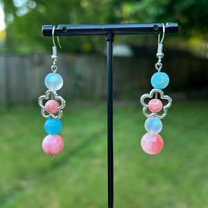 May include: A pair of dangle earrings with a floral design. Each earring features a silver-colored flower charm, pink, blue, and iridescent beads. The earrings are suspended from silver-colored ear wires.
