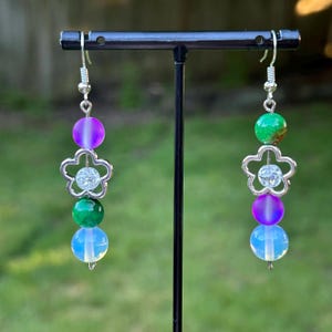 May include: A pair of dangle earrings with a floral design. Each earring features a silver-colored flower charm with a clear gem center, and is adorned with a gradient purple bead, a green bead, and a translucent blue bead. The earrings hang from silver-colored hooks.