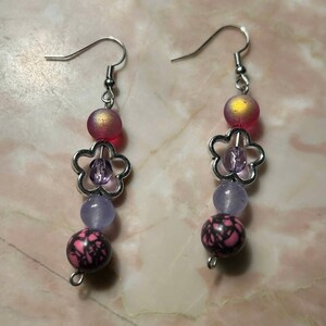 May include: A pair of dangle earrings with a silver flower charm, pink, purple, and iridescent beads.