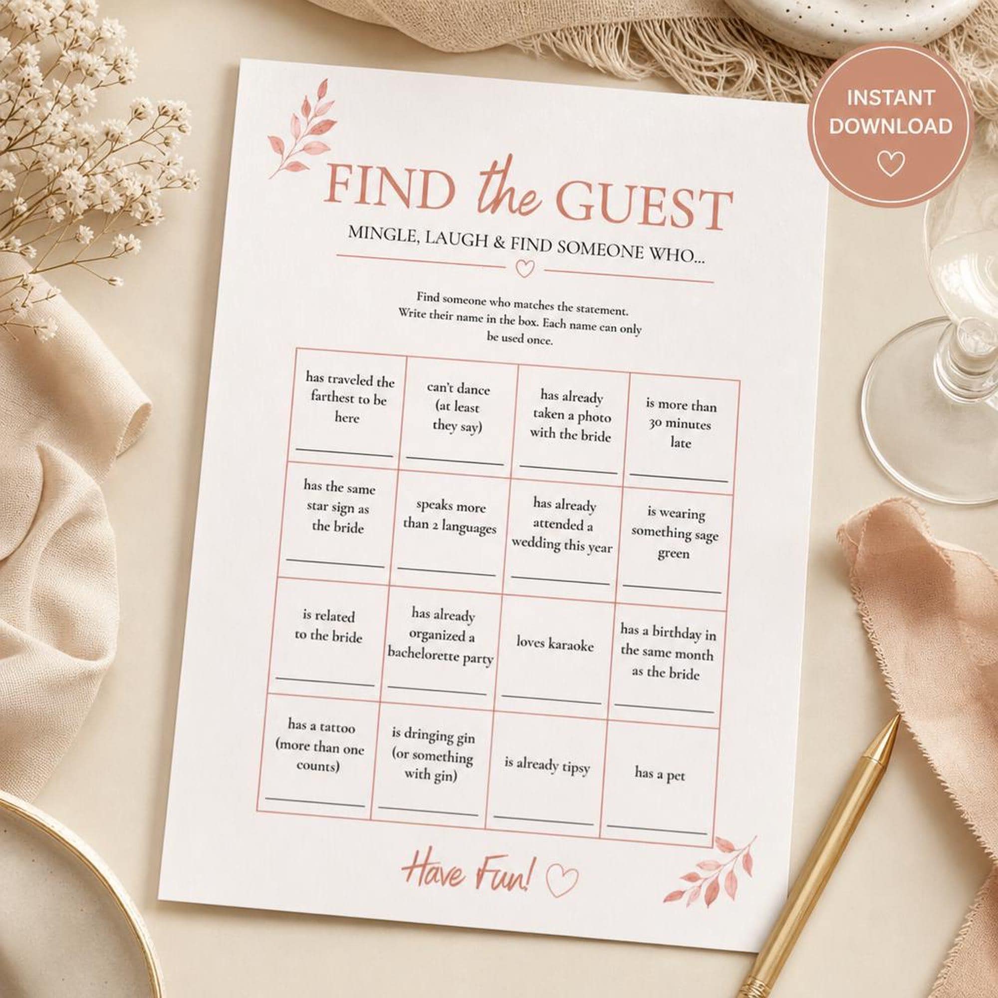 Printable wedding guest bingo game with pink floral accents, displayed on a table beside glassware, fabric, flowers and a pen.