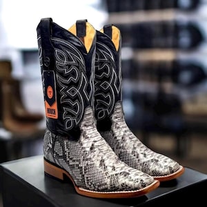 May include: A pair of black and white cowboy boots with a snakeskin pattern. The boots have a black leather shaft with intricate stitching and a brown leather sole. The boots are labeled "Rooch" on the side.