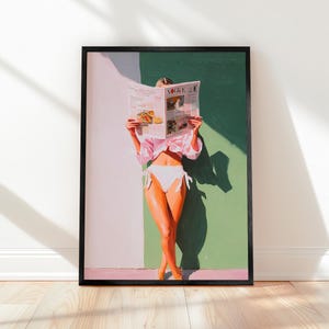 May include: Framed art print featuring a person in a pink shirt and white bikini, holding a newspaper with the word "VOGUE" visible. The artwork has a green and white background, with a shadow cast on the wall. The print is in a black frame.