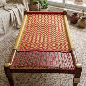 May include: A handcrafted wooden bench with a woven red and gold pattern. The bench has carved details and brass accents on the legs. The design is a blend of traditional craftsmanship and a warm color palette.