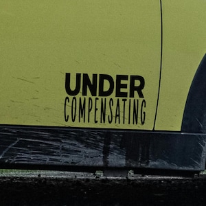 May include: A section of a light green vehicle door with the words "UNDER COMPENSATING" in black, bold font. The door has some scratches and is slightly dirty near the bottom.
