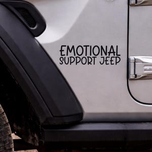 May include: A black decal with the words "EMOTIONAL SUPPORT JEEP" in a bold, sans-serif font is affixed to the side of a silver Jeep. The decal is centered and positioned on the vehicle's door, with the black fender and tire visible.