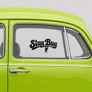 May include: A lime green car window featuring a black vinyl decal with the text Slug Bug in a bold, rounded font. Below the text, a small graphic of a classic car silhouette is visible, centered on the glass surface of the vehicle door.