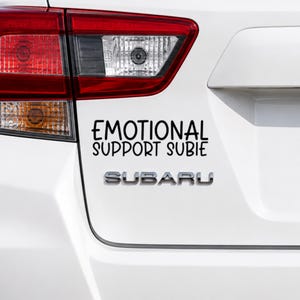 May include: A white car trunk featuring a black vinyl decal with the text EMOTIONAL SUPPORT SUBIE in a playful, rounded font, positioned above the silver metallic Subaru emblem and the red and clear rear taillight assembly of the vehicle.