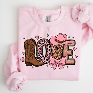 May include: A light pink sweatshirt with a graphic design that says "LOVE". The design includes a brown cowboy boot, a pink cowboy hat, a pink bow, and the letter "O" filled with a leopard print pattern. Small pink hearts and stars surround the word.