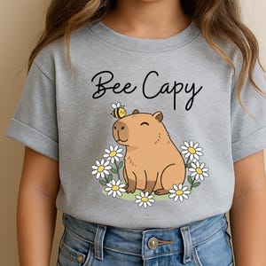 A person wearing a gray t-shirt with a cartoon image of a capybara surrounded by flowers and a bee.