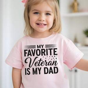 May include: A light pink t-shirt with the text "MY FAVORITE Veteran IS MY DAD" in black. The shirt has a crew neck and short sleeves. The text is centered on the front of the shirt.