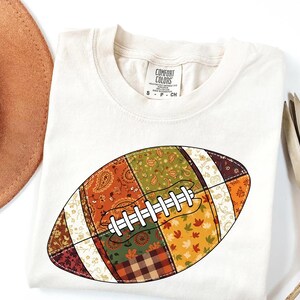 May include: Cream-colored t-shirt featuring a colorful patchwork football design. The football has a white lace design and is made up of various fall-themed patterns in shades of orange, green, and brown. The shirt has a Comfort Colors tag.