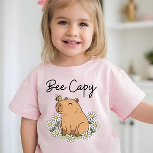 A young girl wearing a pink t-shirt with a cartoon image of a capybara and the text "Bee Cappy" printed on it.