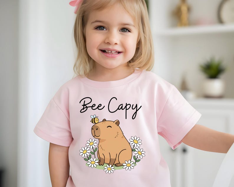 A young girl wearing a pink t-shirt with a cartoon image of a capybara and the text "Bee Cappy" printed on it.