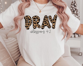 May include: White t-shirt with the word "PRAY" in leopard print lettering. Below the word "PRAY" is the text "colossians 4:2" in a simple black font. The shirt is worn by a person.