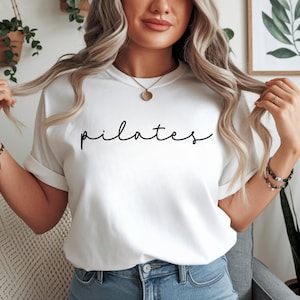 May include: White t-shirt with the word "pilates" in a black script font. The shirt has short sleeves and a crew neck. The model is wearing blue jeans and a gold necklace.