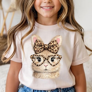 May include: A light pink t-shirt featuring a detailed illustration of a cat wearing heart-shaped glasses and a bow with a leopard print design. The cat's fur is a mix of brown and tan, with pink accents on the ears. The shirt is worn by a young person.