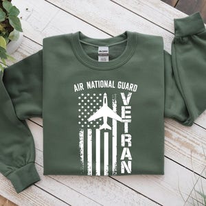 May include: An olive green sweatshirt with the text "AIR NATIONAL GUARD VETERAN" in white. The design features a stylized American flag with a white airplane silhouette. The sweatshirt has long sleeves and a crew neck.