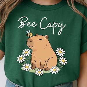 A green t-shirt with a cartoon image of a capybara surrounded by flowers and a bee.