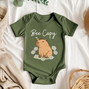 A green baby onesie with a cartoon image of a capybara and flowers, along with the text "Bee Cappy" printed on it, is displayed on a bed with a straw hat and other clothing items.