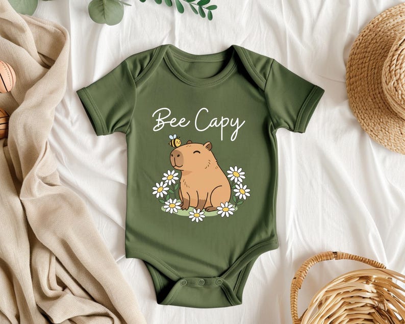 A green baby onesie with a cartoon image of a capybara and flowers, along with the text "Bee Cappy" printed on it, is displayed on a bed with a straw hat and other clothing items.