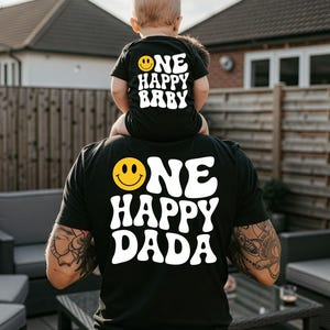 May include: A black short-sleeved t-shirt with white text reading ONE HAPPY DADA and a yellow smiley face, paired with a black baby bodysuit featuring white text reading ONE HAPPY BABY and a yellow smiley face on the back.