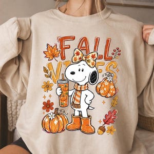 May include: A beige crewneck sweatshirt featuring a graphic of a white cartoon dog wearing a scarf, a bow, and orange boots. The dog holds a tumbler. The design includes the text FALL VIBES, orange pumpkins, autumn leaves, and small flowers.
