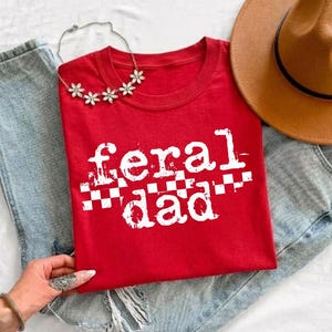 A red t-shirt with the text "feral dad" printed on it, along with a brown hat and a necklace.