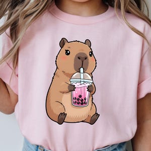 May include: A light pink t-shirt featuring a cartoon capybara drinking a pink bubble tea. The capybara is brown with a light brown belly and is holding a clear cup with a pink drink and black boba pearls.