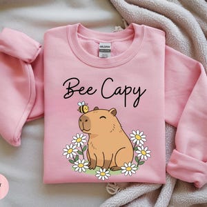 A pink sweatshirt with a cartoon image of a cute brown bear and flowers, along with the text "Bee Cappy" printed on it.