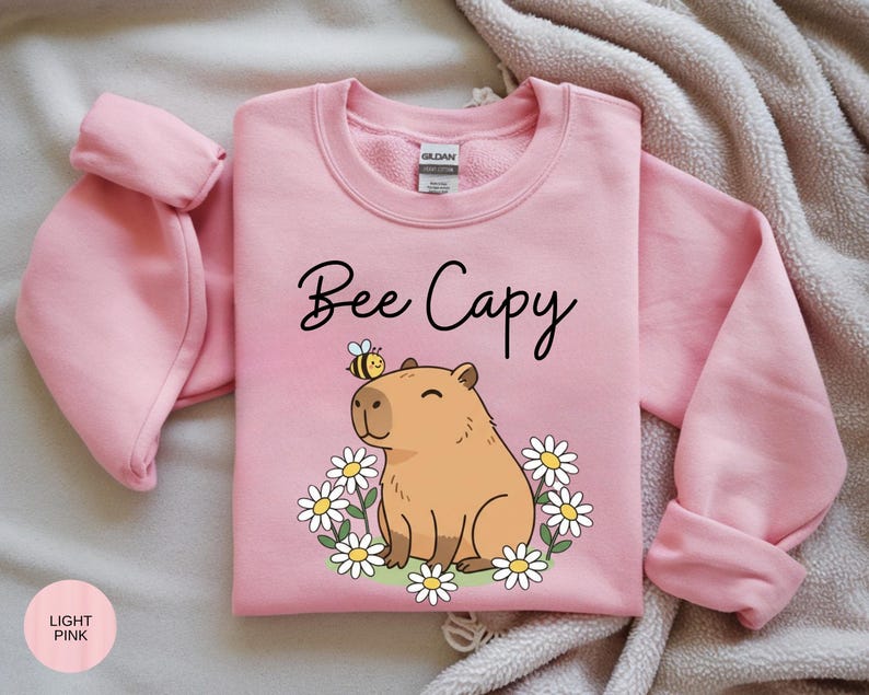 A pink sweatshirt with a cartoon image of a cute brown bear and flowers, along with the text "Bee Cappy" printed on it.