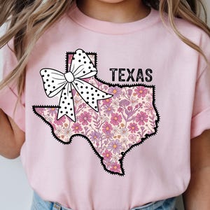 May include: Pink t-shirt featuring a floral Texas outline with a black border. A white bow with black polka dots is above the Texas outline. The word "TEXAS" is printed in black above the state outline.