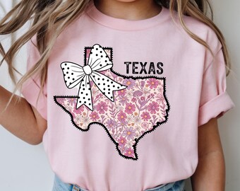 May include: Pale pink t-shirt with a floral Texas outline and a black border. A white bow with black polka dots is above the Texas outline. The word "TEXAS" is printed in black above the state outline.