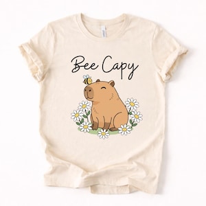 A white t-shirt with a cartoon image of a capybara surrounded by flowers and a bee.