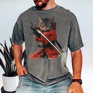 May include: Grey t-shirt with a graphic of a cat in a samurai outfit, complete with a red robe and swords. The cat has a determined look. The shirt has a vintage wash.