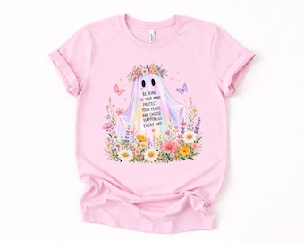 May include: A light pink t-shirt featuring a central graphic of a ghost wearing a flower crown, surrounded by colorful wildflowers and butterflies. The ghost has text that reads: BE KIND TO YOUR MIND, PROTECT YOUR PEACE, AND CHOOSE HAPPINESS EVERY DAY.