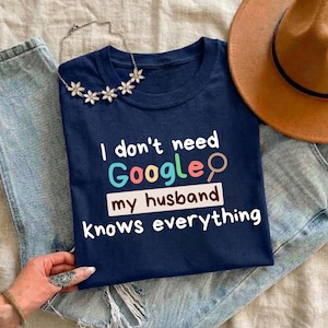 May include: Navy blue t-shirt with the text "I don't need Google my husband knows everything" in white, blue, green, and red. A brown hat and a silver necklace with flower-shaped pendants are also visible.