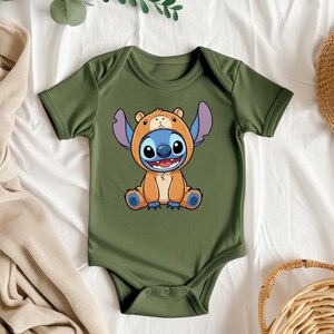 A green baby onesie with a cartoon character design is displayed on a bed, surrounded by various clothing items and accessories.