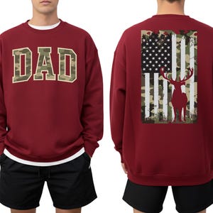 May include: A burgundy sweatshirt with the word "DAD" in a camouflage design across the chest. The back of the sweatshirt features an American flag design with a deer silhouette. The sweatshirt has a crew neck and long sleeves.