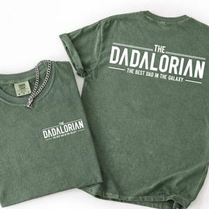 May include: A sage green t-shirt featuring white text on the front left chest and a larger version on the back. The text reads THE DADALORIAN THE BEST DAD IN THE GALAXY. A silver-toned metal chain necklace rests on the collar of the folded shirt.