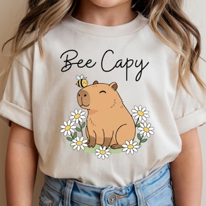 May include: Beige t-shirt with the text "Bee Capy" in black script. The shirt features a cartoon capybara with a bee on its head, surrounded by white daisies with yellow centers.